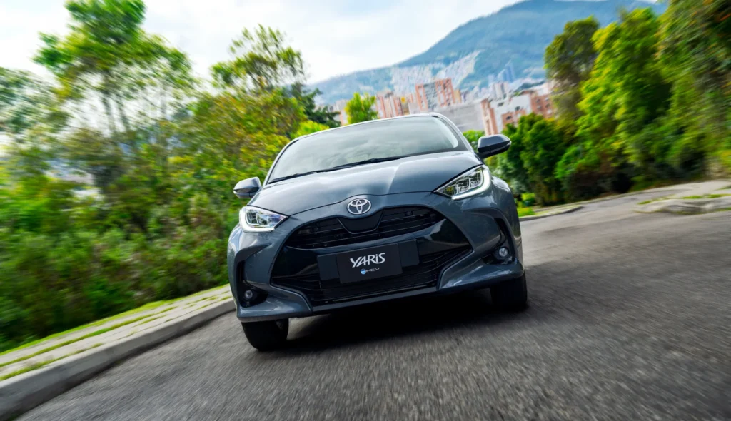Toyota Yaris HEV de frente en carretera, con la ciudad al fondo